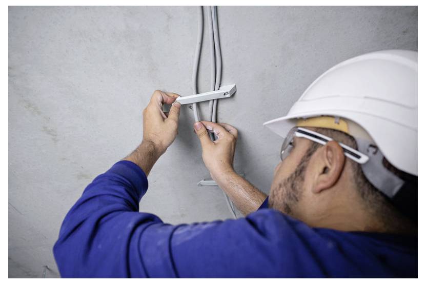 Osoba w kasku ochronnym i okularach ochronnych instaluje przewody elektryczne na ścianie, używając narzędzia do mocowania metalowych uchwytów.