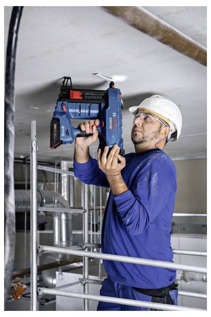 Robotnik budowlany w niebieskim mundurze i białym kasku ochronnym, używający elektronarzędzia do instalacji przewodów elektrycznych lub rur na suficie na terenie budowy.