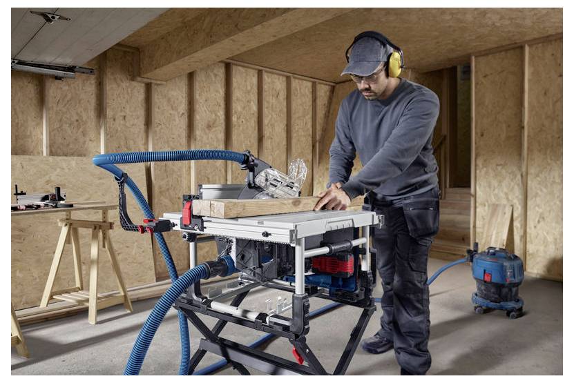 Mężczyzna używający piły elektrycznej w warsztacie stolarski, ubrany w sprzęt ochronny taki jak rękawice i ochronniki słuchu. Wąż odkurzacza podłączony do kontroli pyłu.