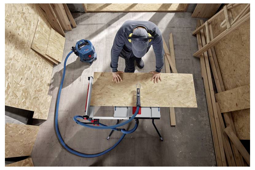 Osoba w czapce i nausznikach tnie drewnianą deskę na warsztacie z podłączonym odkurzaczem, wokół której znajdują się drewniane panele.