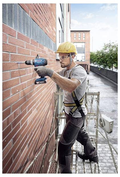 Robotnik budowlany w żółtym kasku i szelkach bezpieczeństwa używa wiertarki elektrycznej na murowanej ścianie ze scaffoldingu na zewnątrz budynku.