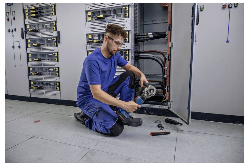 Technik w niebieskim ubraniu roboczym klęczy na podłodze wyłożonej kafelkami, używając ręcznego narzędzia przed otwartą rozdzielnicą elektryczną z różnymi kablami.