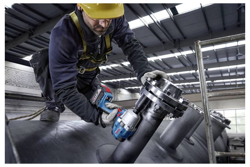 Pracownik w odzieży ochronnej i żółtym kasku używa elektronarzędzia do dokręcania śrub na dużej przemysłowej rurze wewnątrz fabryki.