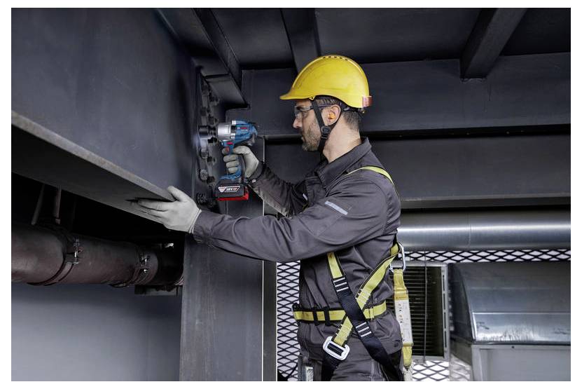 Pracownik w sprzęcie ochronnym, w tym żółtym kasku i szelkach bezpieczeństwa, używa narzędzia elektrycznego do zamocowania metalowych belek w środowisku przemysłowym.