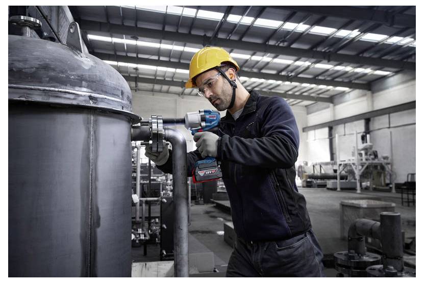 Pracownik w odzieży ochronnej obsługuje elektronarzędzie na rurociągach przemysłowych wewnątrz dużej fabryki, podkreślając koncentrację na konserwacji przemysłowej.