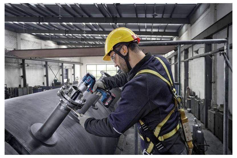 Pracownik w kamizelce bezpieczeństwa i kasku używa elektronarzędzia na dużej metalowej konstrukcji w środowisku fabrycznym.