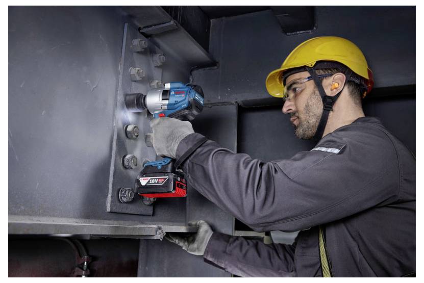Pracownik używający bezprzewodowego klucza udarowego do dokręcania śrub na dużej metalowej konstrukcji, ubrany w kask ochronny i okulary ochronne.