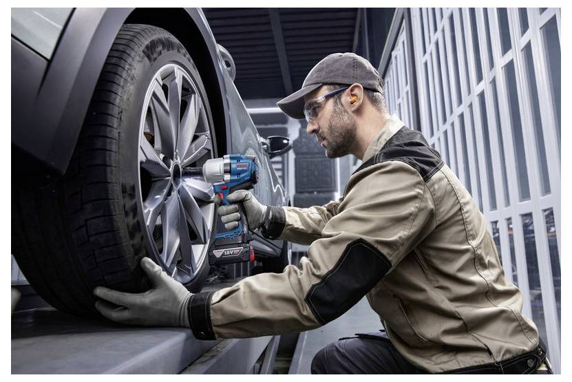 Mechanik w rękawicach i sprzęcie ochronnym używa elektrycznego klucza do dokręcania śrub na kole samochodu wewnątrz warsztatu.