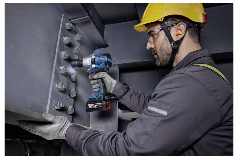 Robotnik budowlany w środkach ochrony indywidualnej, w tym żółtym kasku ochronnym i rękawicach, używający elektronarzędzia do dokręcania śrub na dużej metalowej konstrukcji.