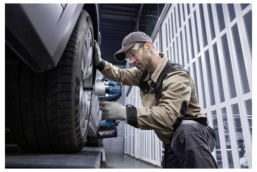 Mechanik w odzieży ochronnej używa elektronarzędzia do dokręcenia koła samochodu w warsztacie, podkreślając konserwację i bezpieczeństwo pojazdu.