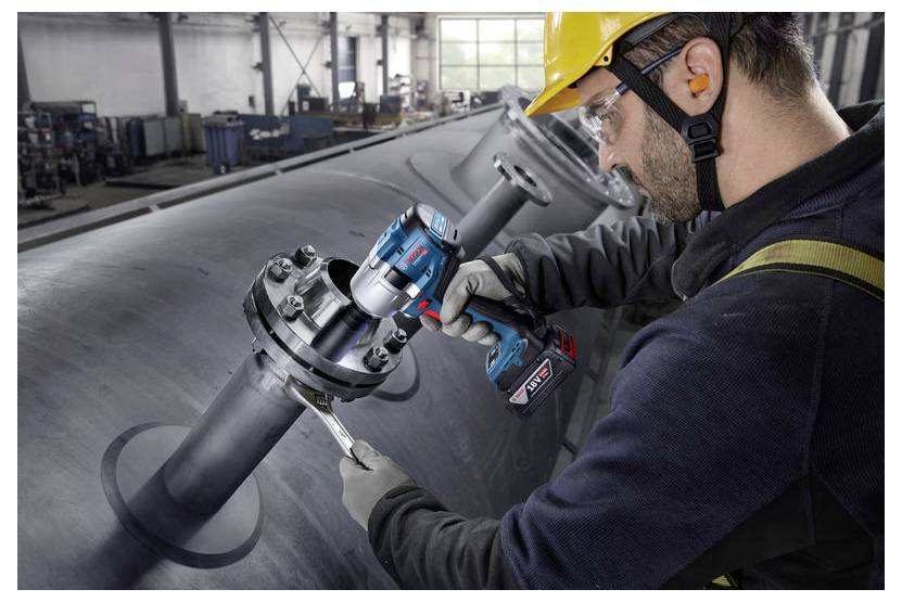 Pracownik w ubraniu ochronnym używa elektronarzędzia do dokręcania śruby na dużej przemysłowej rurze wewnątrz hali fabrycznej.