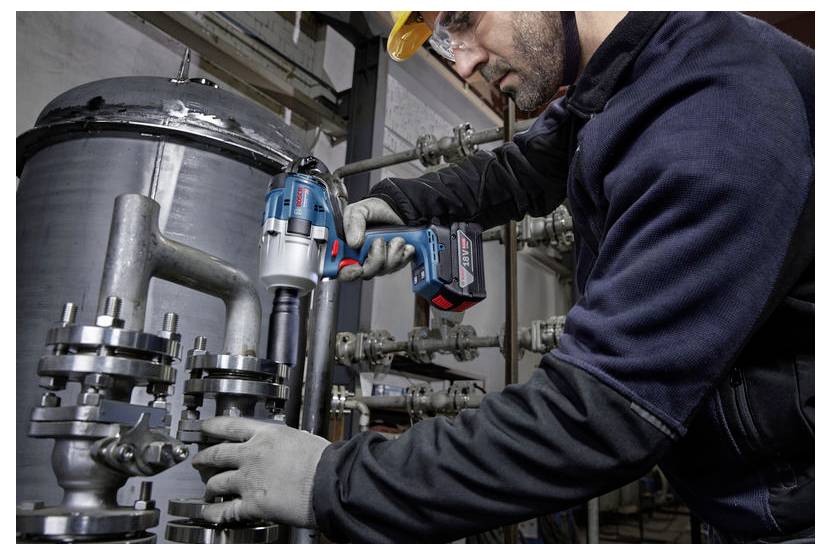Osoba w odzieży ochronnej używa elektronarzędzia na przemysłowych rurach metalowych, co wskazuje na trwające prace konserwacyjne lub naprawcze.