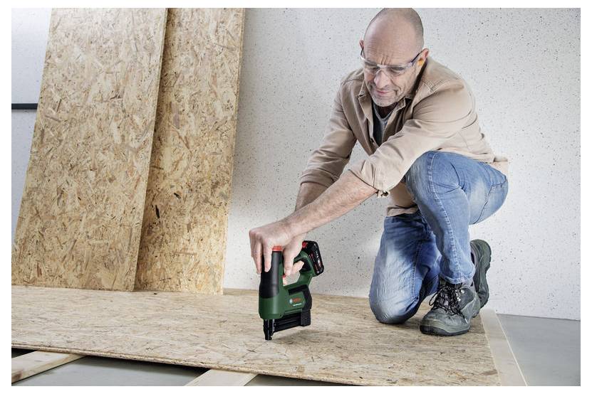 Mężczyzna używający zielonego narzędzia elektrycznego na drewnie, klęczący w ochronnych okularach, sugerujący prace stolarskie lub działalność budowlaną.