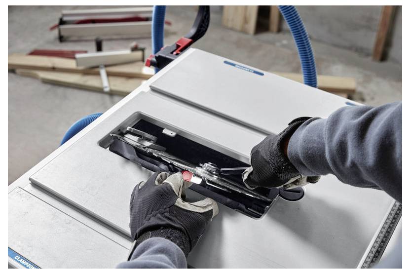 Osoba w rękawicach reguluje ostrze piły stołowej z podłączonym niebieskim wężem. Na tle widoczne są kawałki drewna i narzędzie pomiarowe.