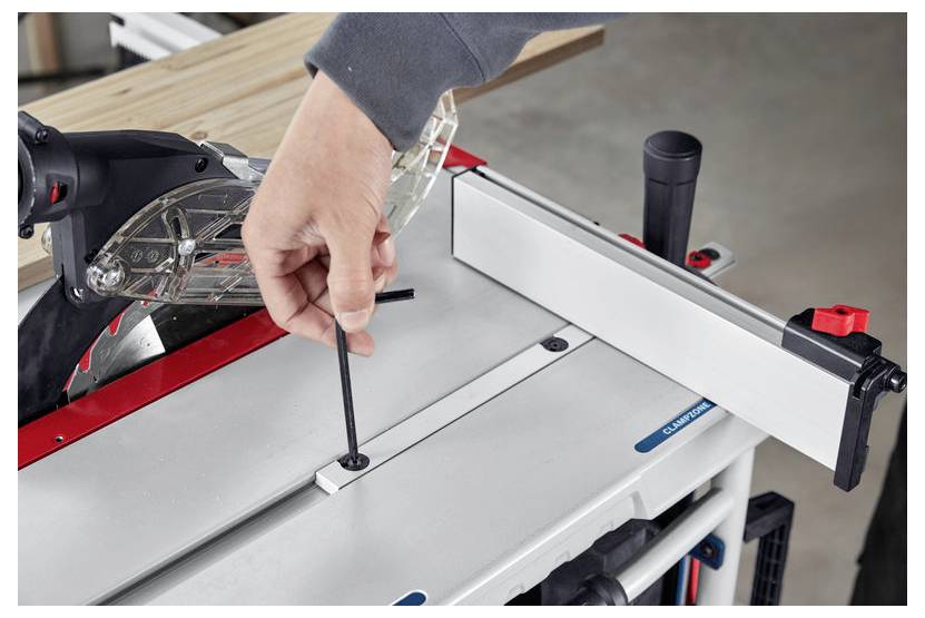 Osoba reguluje ustawienie brzeszczotu piły stołowej za pomocą klucza sześciokątnego. Drewniana deska jest położona na powierzchni piły.