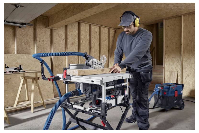 Osoba w odzieży ochronnej obsługująca piłę stołową na desce drewnianej w warsztacie, z podłączonym sprzętem do odpylania.
