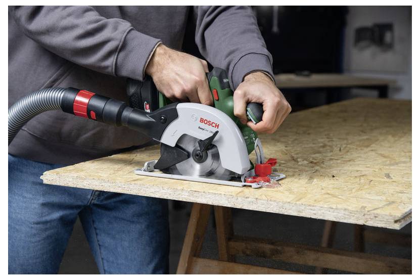 Osoba używa piły tarczowej Bosch do cięcia arkusza drewna na stole warsztatowym. Pył jest zbierany przez podłączony wąż odkurzacza.