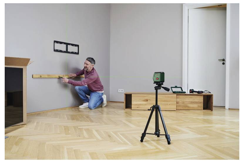 Mężczyzna klęczący na podłodze używa poziomicy do pomiaru ściany pod uchwyt telewizora. Statyw z laserowym poziomem stoi na drewnianej podłodze.