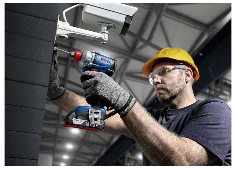 Pracownik w kasku ochronnym używający niebiesko-czerwonej wiertarki do zamontowania białej kamery bezpieczeństwa na ścienie.