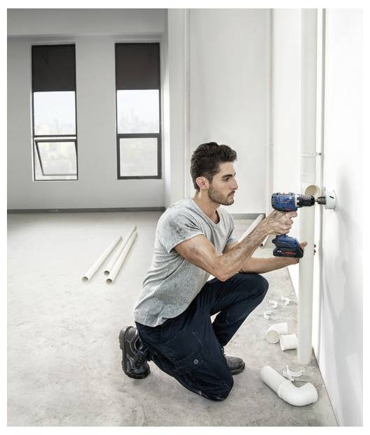 Osoba klęczy na podłodze wewnątrz pomieszczenia, używając wiertarki elektrycznej do montażu rur wzdłuż ściany. Różne rury są rozrzucone dookoła.