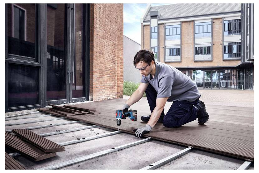 Osoba ubrana w sprzęt ochronny montuje drewniane deski na tarasie na świeżym powietrzu, używając elektronarzędzia, z nowoczesnym budynkiem w tle.