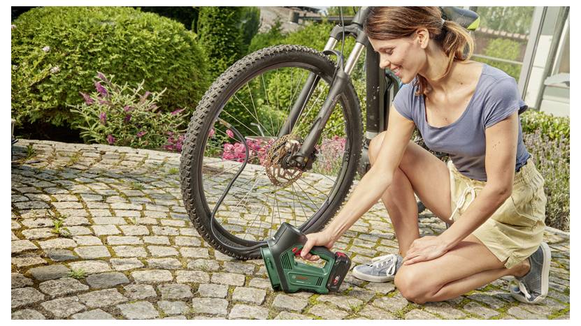 Osoba ubrana w niebieską koszulkę i beżowe szorty kuca na kamiennej ścieżce, używając zielonego przenośnego urządzenia do pompowania opony roweru.