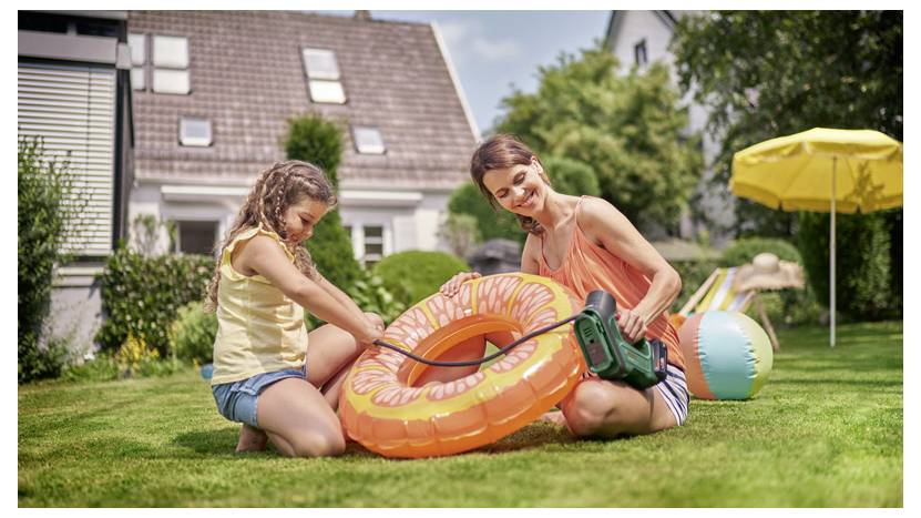 Kobieta i dziewczynka siedzą na trawie w ogrodzie, pompując dużą, pomarańczową dmuchaną oponę basenową za pomocą pompki, a w tle widać domy.