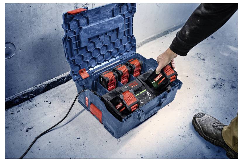 Osoba umieszcza akumulator elektronarzędzia w kolorze czerwonym i czarnym w niebieskiej ładowarce z wieloma gniazdami na betonowej podłodze.