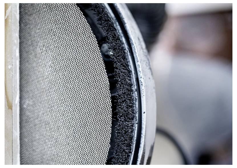 Zbliżenie na teksturowaną metalową siatkę z okrągłą, gęstą szczotką z czarnych włosków na krawędzi, prawdopodobnie będącą częścią mechanizmu czyszczącego lub filtracyjnego.