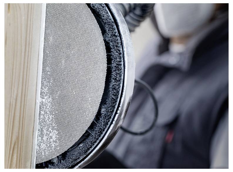 Osoba ubrana w odzież ochronną używa szlifierki elektrycznej na drewnianej powierzchni, podkreślając postęp prac stolarskich lub ciesielskich.