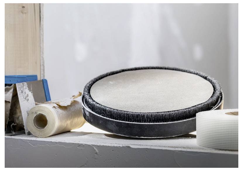 Podkładka do ćwiczeń perkusyjnych spoczywa na półce obok rolki taśmy i niebiesko obramowanego narzędzia, sugerując scenę związaną z praktyką muzyczną lub pracą naprawczą.