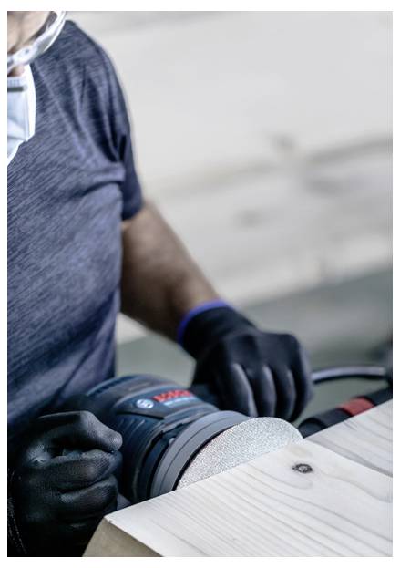 Osoba używająca szlifierki elektrycznej do wygładzania drewnianej deski, nosząca rękawice i maskę ochronną, skupiona na zadaniu, z rozmytym tłem.