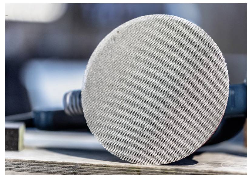 Zbliżenie tarczy papieru ściernego przymocowanej do narzędzia, spoczywającej na drewnianej powierzchni, z rozmytym tłem w tle.