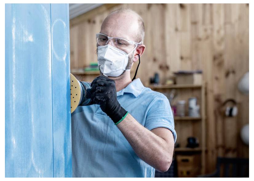 Osoba nosząca okulary ochronne, maskę i rękawice używa szlifierki elektrycznej na niebieskiej drewnianej powierzchni, co sugeruje prace stolarskie lub renowację mebli.