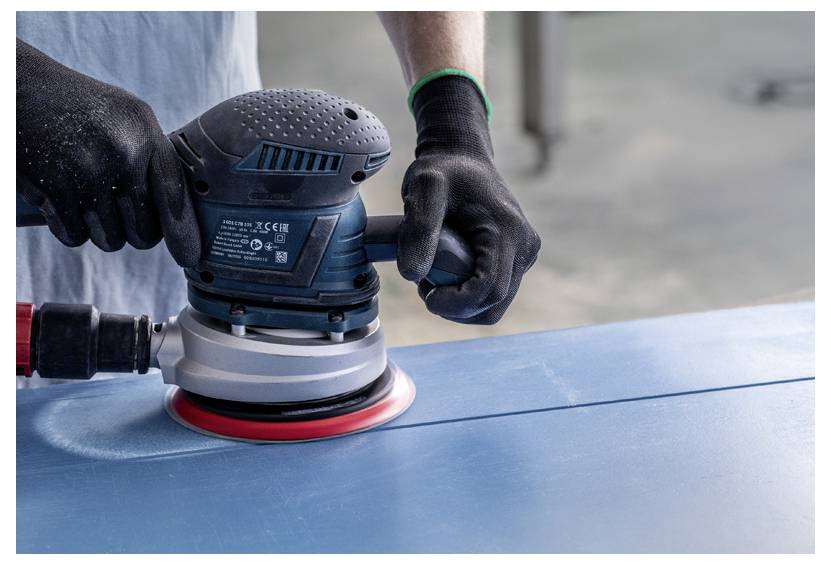 Osoba używa elektrycznej szlifierki na niebieskiej powierzchni, wygładzając ją, podczas gdy nosi czarne rękawice ochronne.