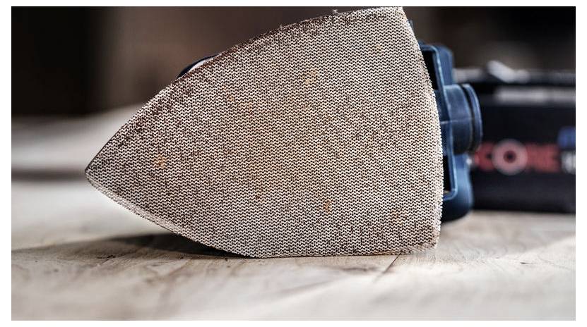 Szczegółowy obraz ręcznej szlifierki z teksturowaną prostokątną nakładką do szlifowania, spoczywającej na drewnianej powierzchni, prezentujący jej ziarnistość i design.