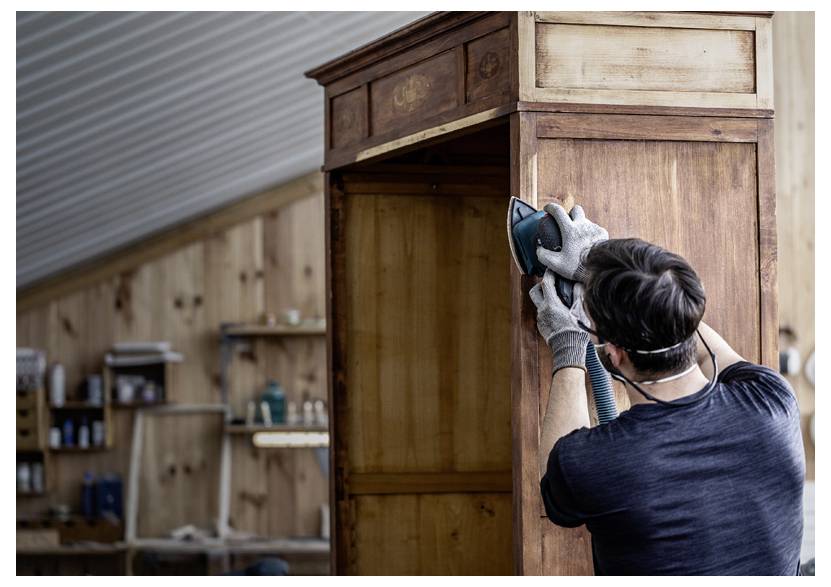 Osoba szlifuje drewniany kredens w warsztacie, nosząc rękawice i maskę, a w tle widać półki i drewniane ściany.