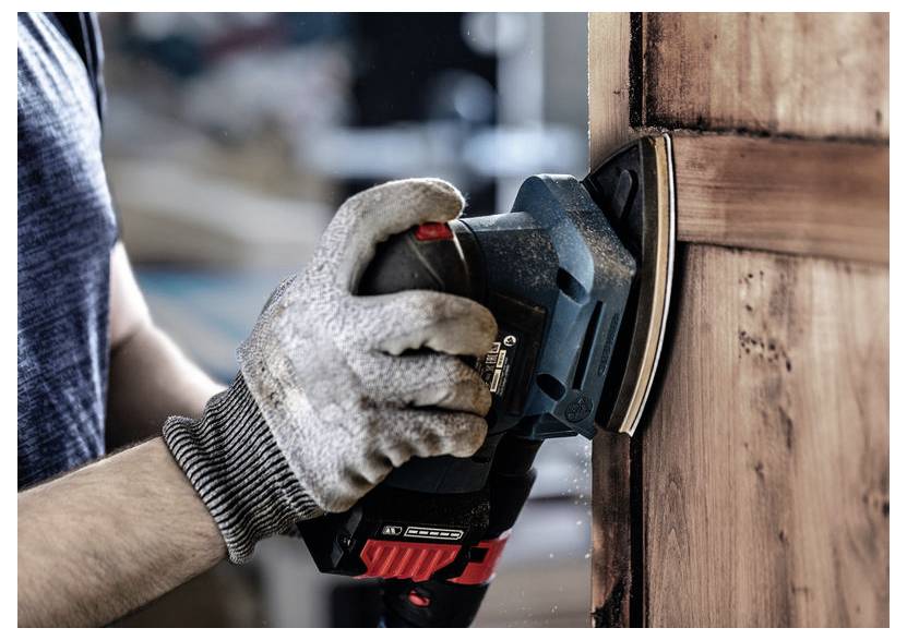 Osoba używająca ręcznej szlifierki do wygładzania powierzchni drewnianych drzwi. Dłoń w rękawicy trzymająca szlifierkę, widoczny pył drewniany.