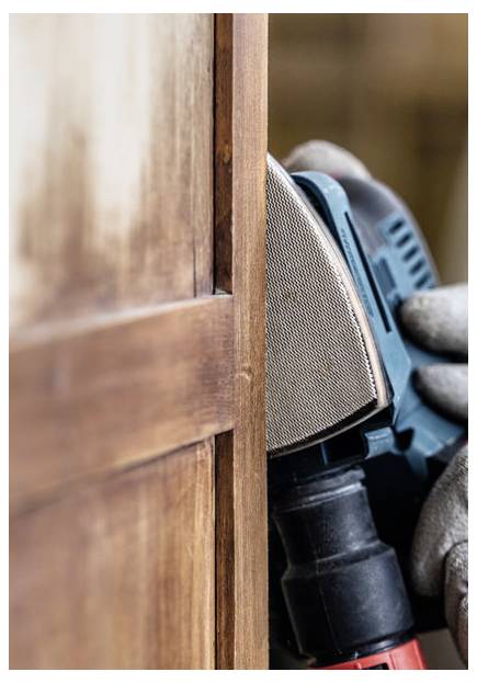 Osoba używa szlifierki elektrycznej na drewnianych drzwiach, wygładzając powierzchnię. Rękawicą zakryte dłonie pewnie trzymają narzędzie, koncentrując się na krawędzi.