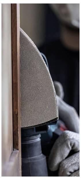 Osoba w rękawicach używa szlifierki elektrycznej na drewnianej powierzchni, koncentrując się na krawędzi, co sugeruje stolarkę lub renowację mebli.