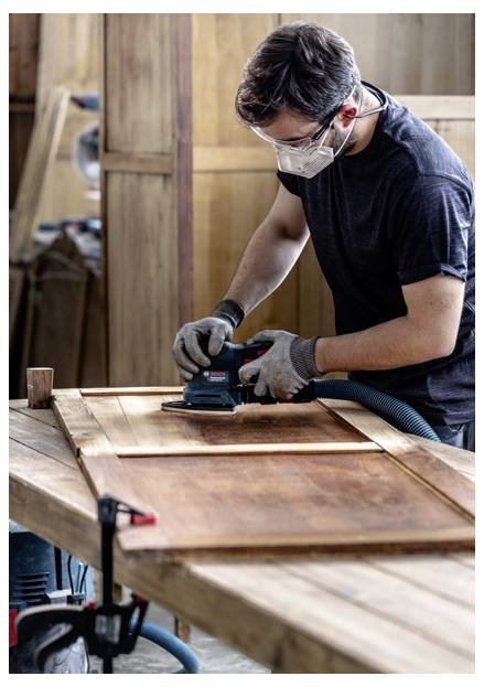 Osoba w okularach ochronnych i masce szlifuje drewniane drzwi na stole warsztatowym w warsztacie stolarski, koncentrując się na górnej powierzchni.