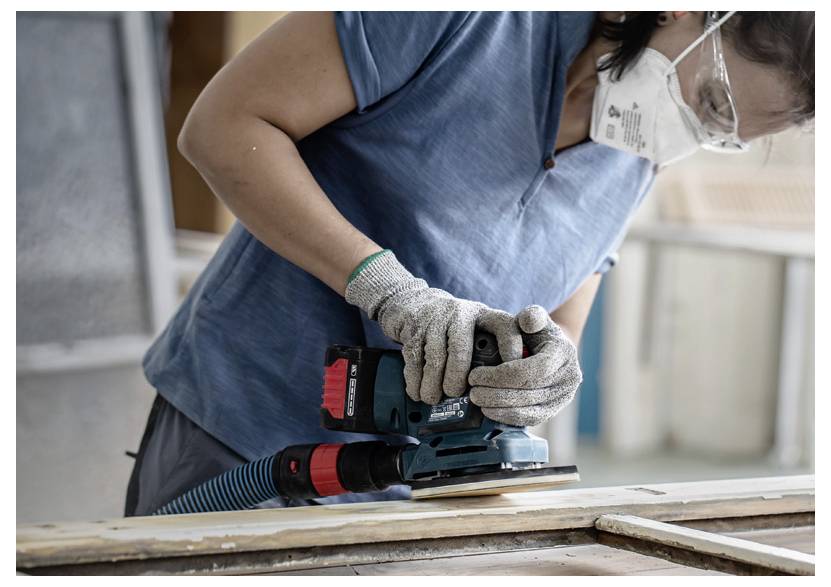 Osoba w masce i rękawicach używa szlifierki elektrycznej na drewnianej powierzchni w środowisku warsztatu.