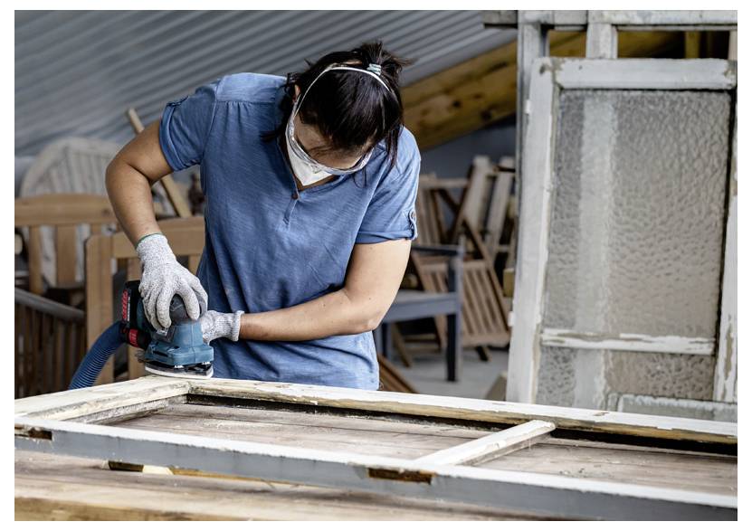 Osoba używająca szlifierki elektrycznej na drewnianych drzwiach w warsztacie, nosząca okulary ochronne, maskę i rękawice ochronne.
