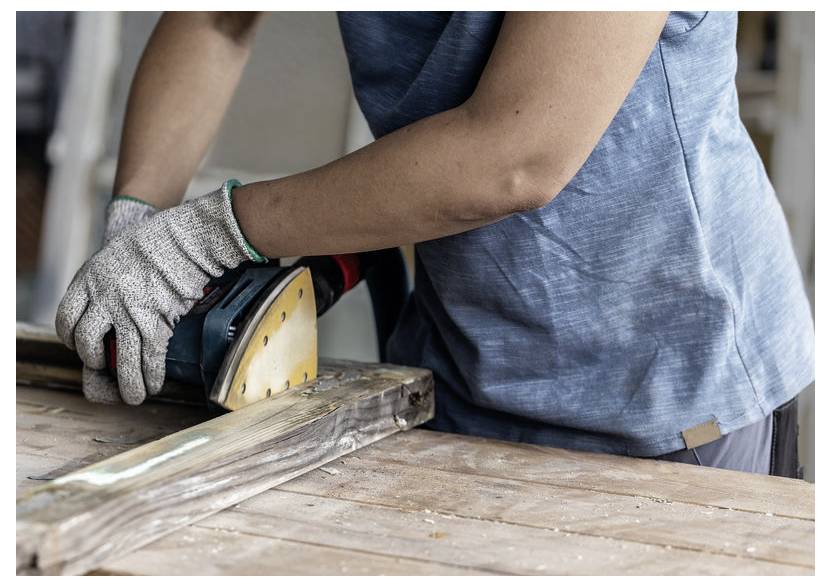 Osoba w rękawicach używa szlifierki elektrycznej do szlifowania drewnianej deski, prezentując stolarkę w warsztacie.