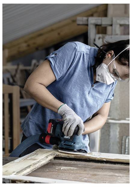 Osoba w niebieskiej koszuli i maseczce ochronnej szlifuje drewnianą powierzchnię szlifierką elektryczną w warsztacie.