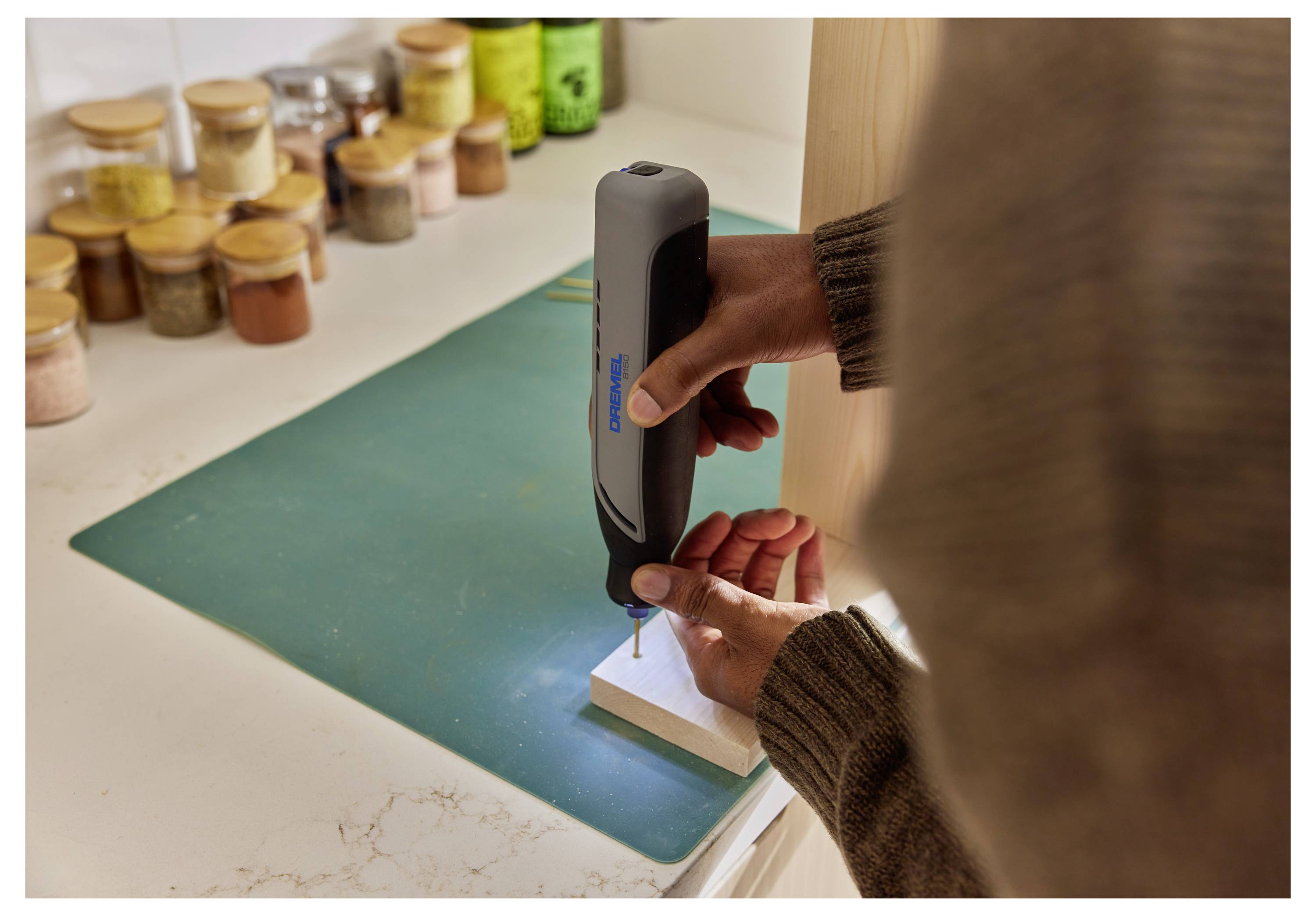 Osoba używająca małej, ręcznej wiertarki elektrycznej na kawałku drewna. W tle na blacie widoczne są różne słoiczki z przyprawami.