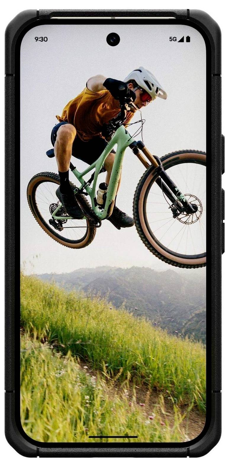 Osoba w kasku wykonuje skok na rowerze górskim na tle wzgórz i czystego nieba, prezentując dynamiczną akcję i sport.