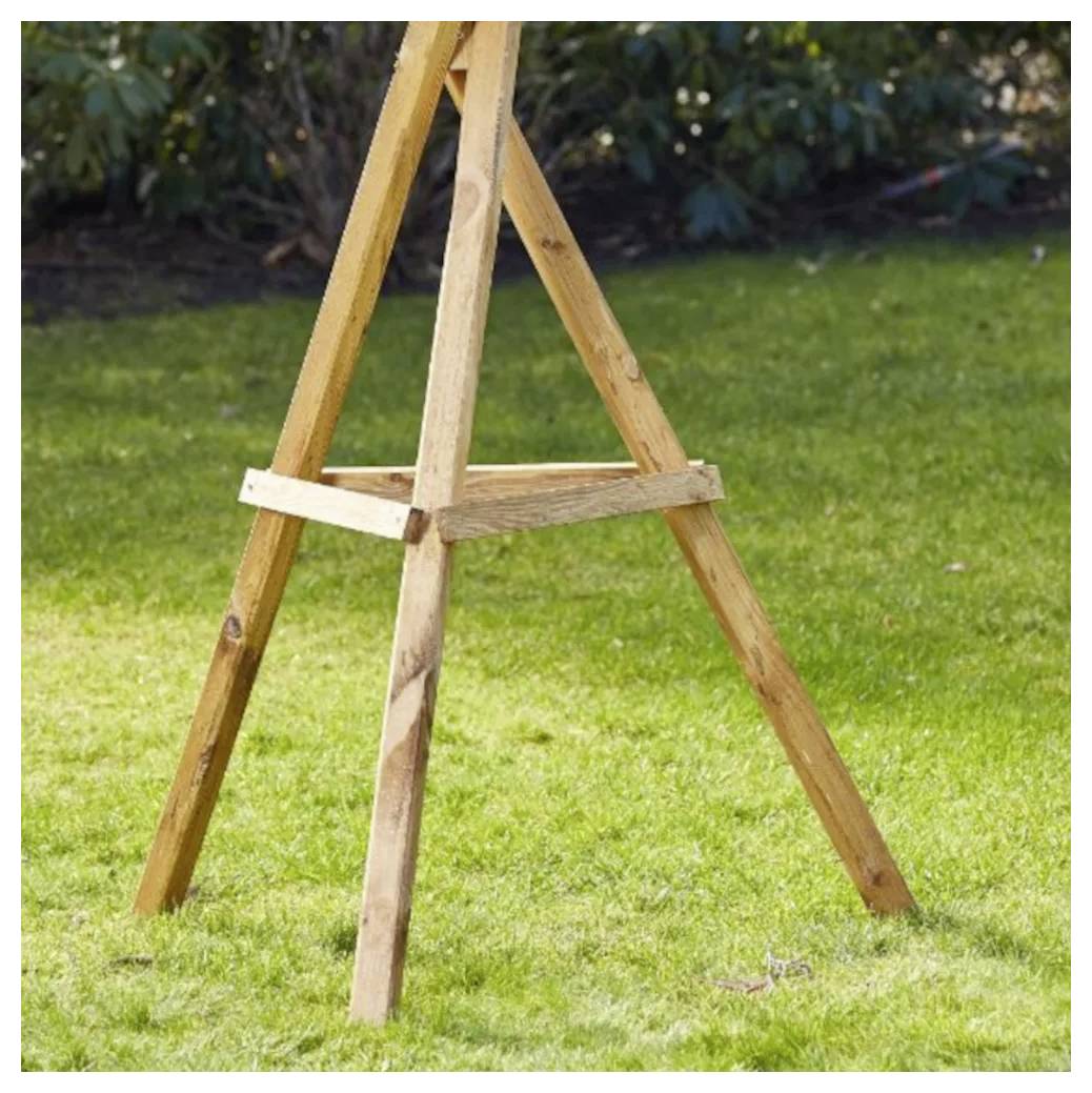 Drewniany sztalugi stojący na zielonej trawie w ogrodowym otoczeniu, z liściastymi krzewami w tle.