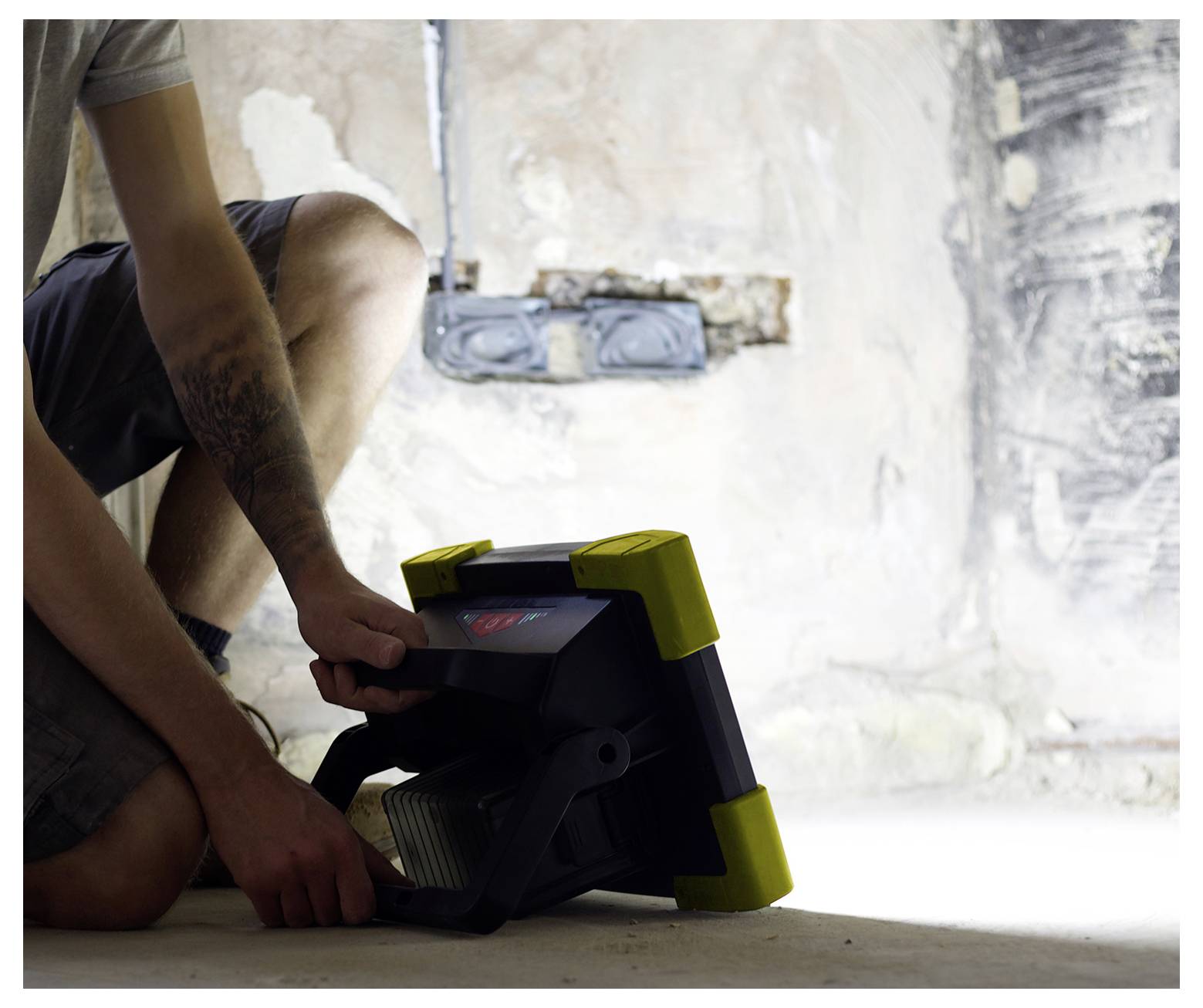 Osoba klęczy na jasnej podłodze, regulując duży przenośny reflektor roboczy w słabo oświetlonym, niedokończonym pomieszczeniu z widocznymi ścianami.