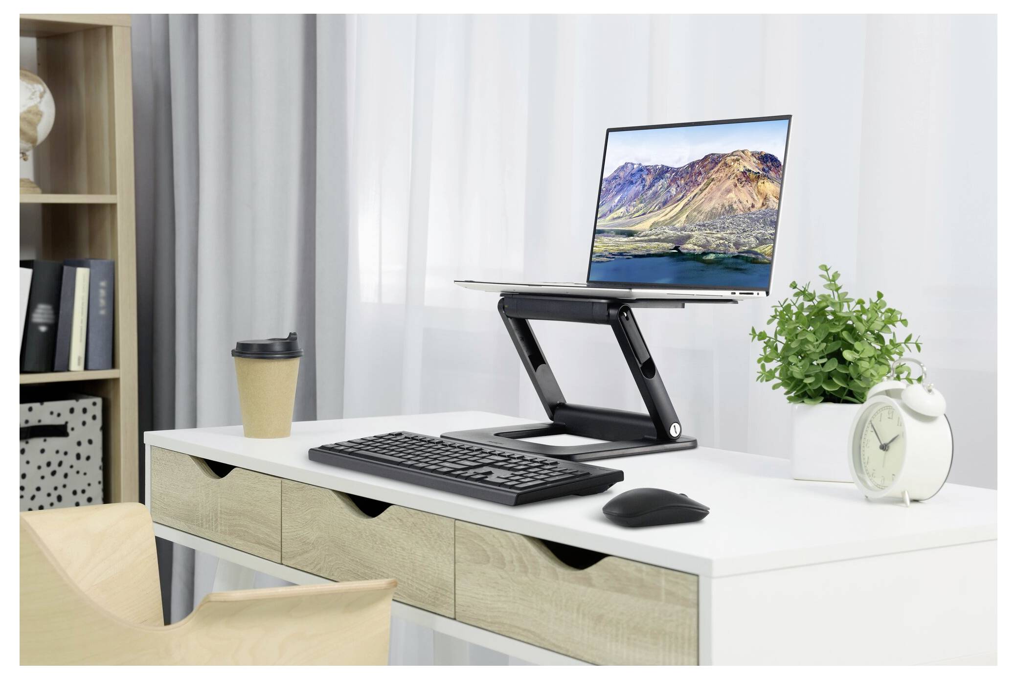 Nowoczesne stanowisko pracy z laptopem na regulowanym statywie, klawiaturą, myszką, kubkiem kawy, małą rośliną i zegarem w jasnym pomieszczeniu.